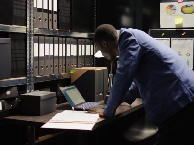 In neatly organised office, african american detective examines evidence with intention of solving crimes. Committed police officer walks up to his desk carrying case file for criminal investigation.
